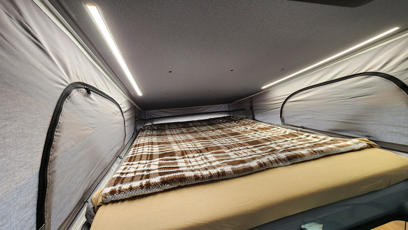 RV rental's cozy pop-top bed features a brown plaid blanket, beige sheets, and bright LED lighting for comfortable camping.