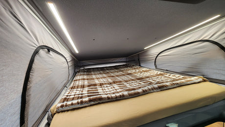RV rental's cozy pop-top bed features a brown plaid blanket, beige sheets, and bright LED lighting for comfortable camping.