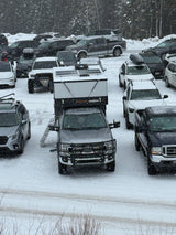 A snowy parking lot fills with cars and an RV rental, a truck camper mounted on a pickup, during heavy winter snowfall.