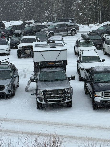 A snowy parking lot fills with cars and an RV rental, a truck camper mounted on a pickup, during heavy winter snowfall.