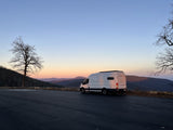 A white rv rental camper van overlooks a tranquil mountain range during a soft sunset, framed by bare trees on a paved lot.