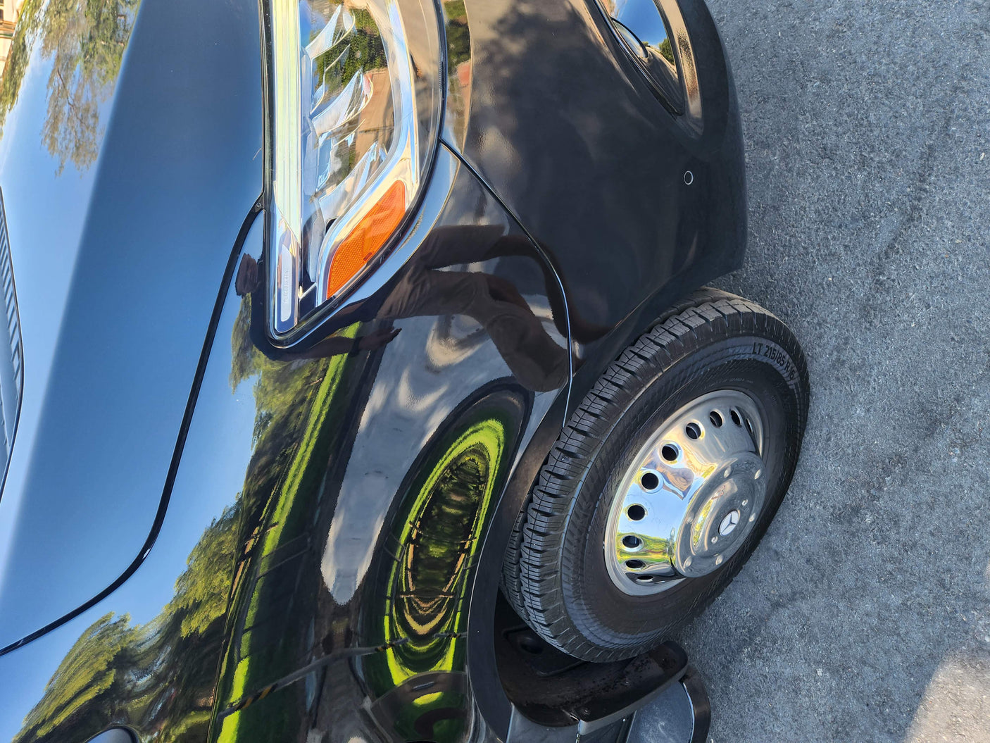 This black Mercedes-Benz van, a potential rv rental, features a sharp headlight and shiny wheel, reflecting trees and sky.