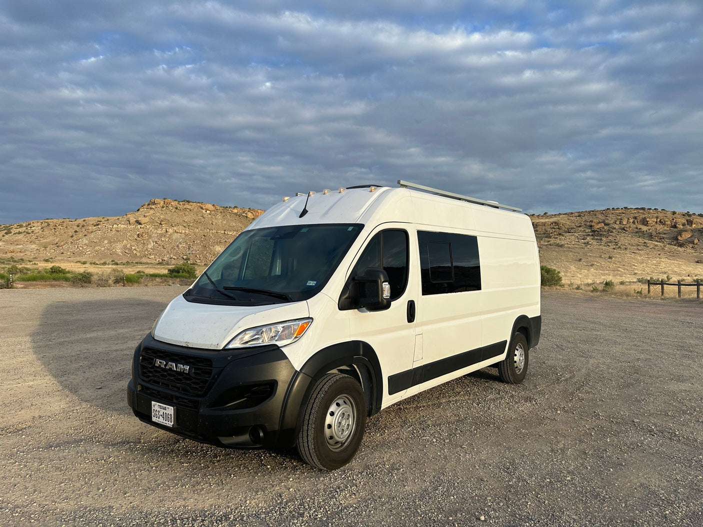 White Ram ProMaster camper van, an rv rental ready for adventure, parked on a gravel lot with rocky desert hills.