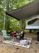 Family enjoying an outdoor meal under an RV rental awning with their dog at a peaceful campground.