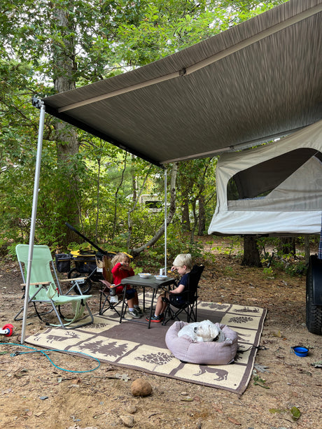 Family enjoying an outdoor meal under an RV rental awning with their dog at a peaceful campground.