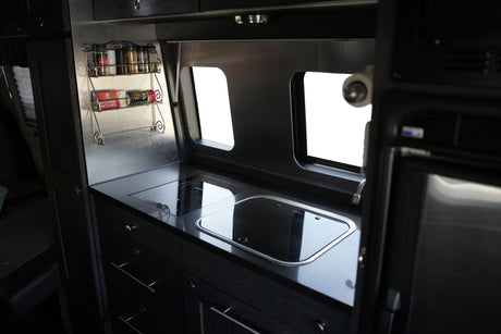 Modern RV rental kitchen with black induction cooktop, sink, and a stainless steel spice rack. Bright windows illuminate the compact galley.