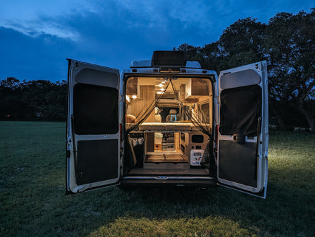 Illuminated RV rental at dusk, with open back doors revealing its compact, cozy interior featuring a bed. Parked on grass.