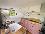 RV rental van interior: Features a cozy bed, white overhead storage, and a functional kitchen with a sink & pink cabinets.