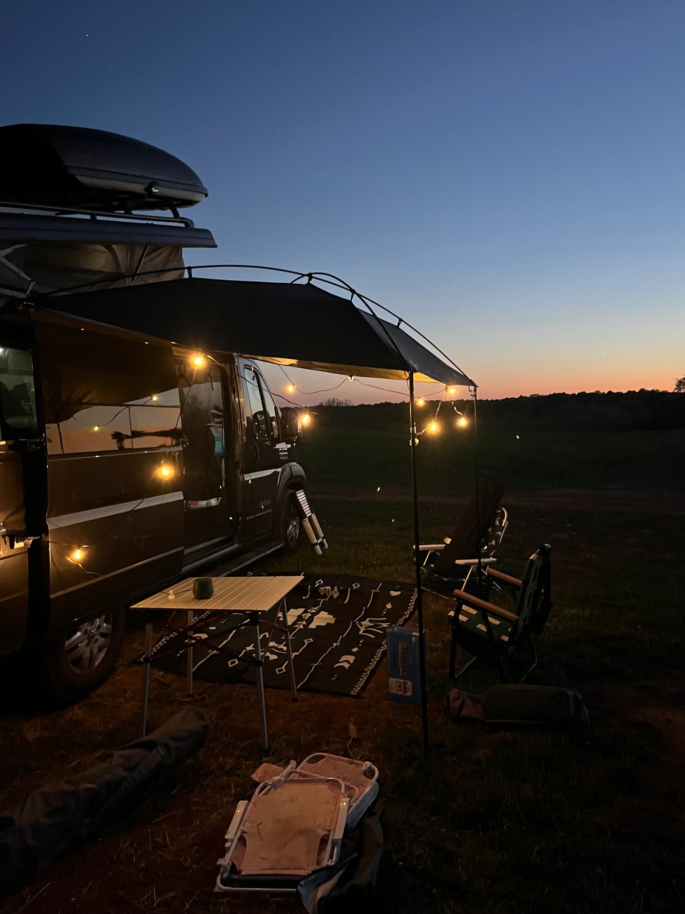 Cozy RV rental setup at dusk: camper van with illuminated awning, table, chairs, and rug on grass under twilight sky.