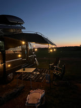 Cozy RV rental setup at dusk: camper van with illuminated awning, table, chairs, and rug on grass under twilight sky.