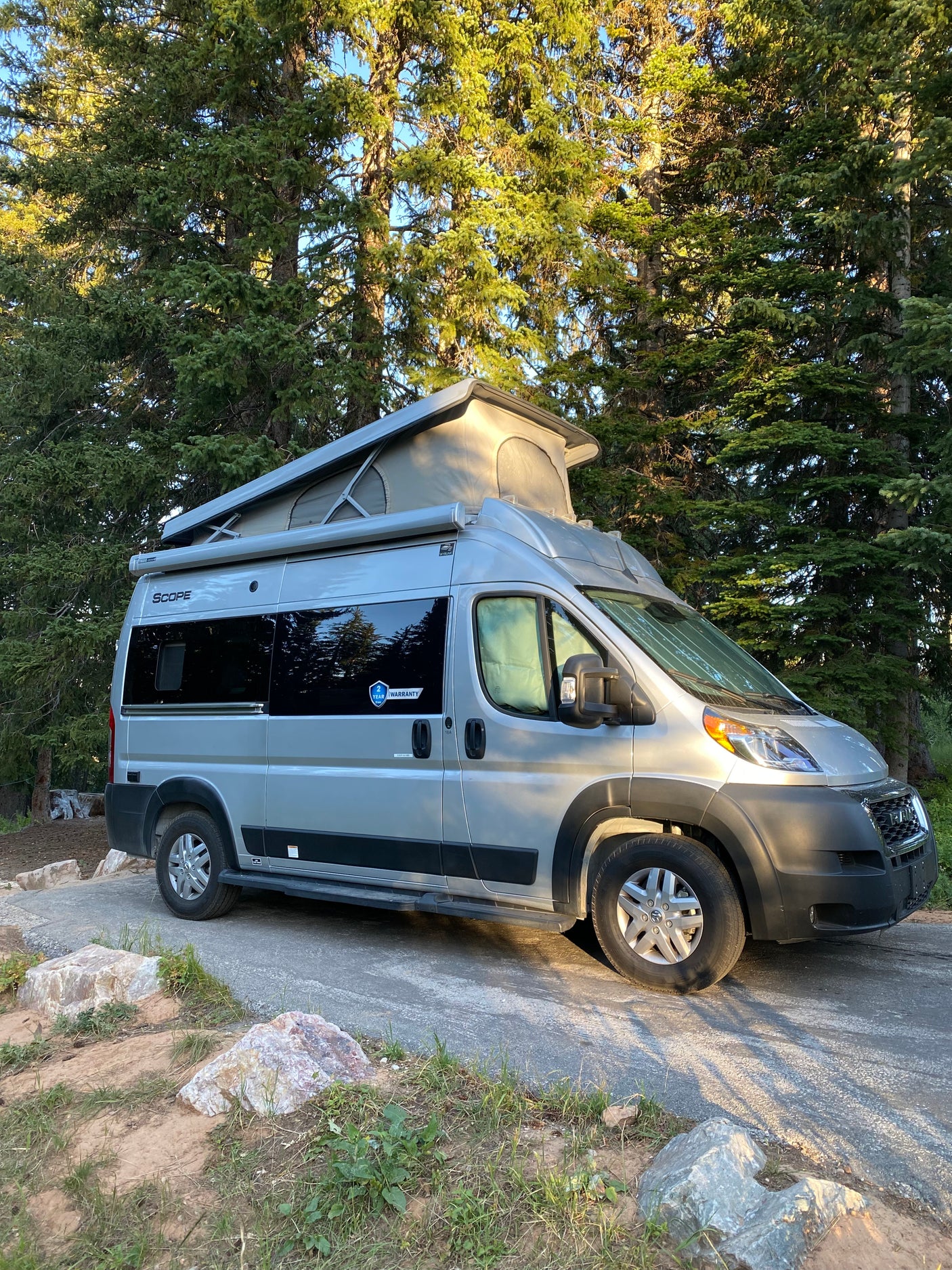 A silver pop-top RV rental camper van is parked on a paved spot among tall evergreen trees, perfect for camping.