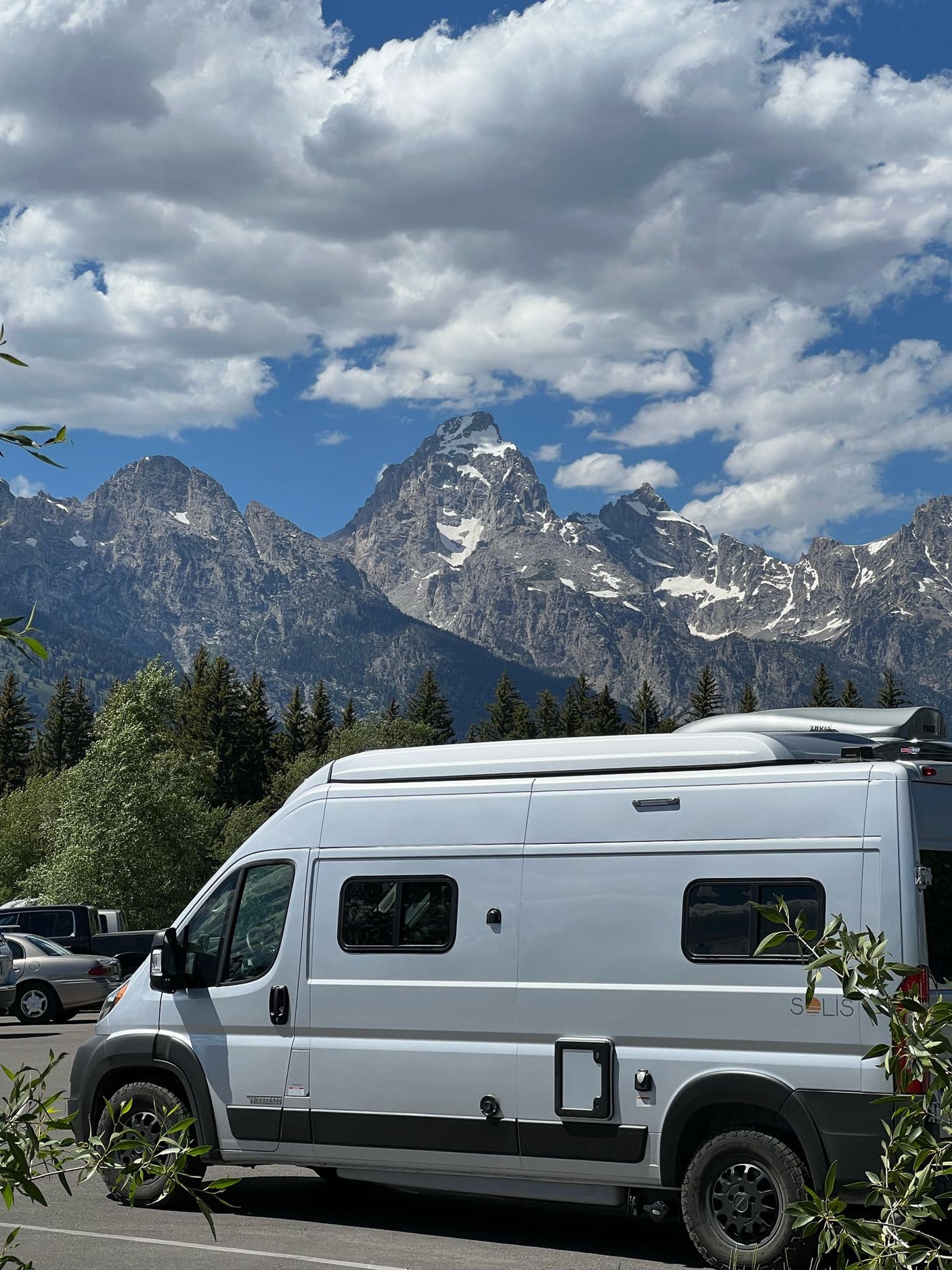 Your next RV rental adventure awaits! A white camper van with stunning Teton mountain range views under a blue, cloudy sky.