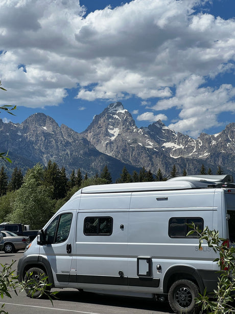 Your next RV rental adventure awaits! A white camper van with stunning Teton mountain range views under a blue, cloudy sky.