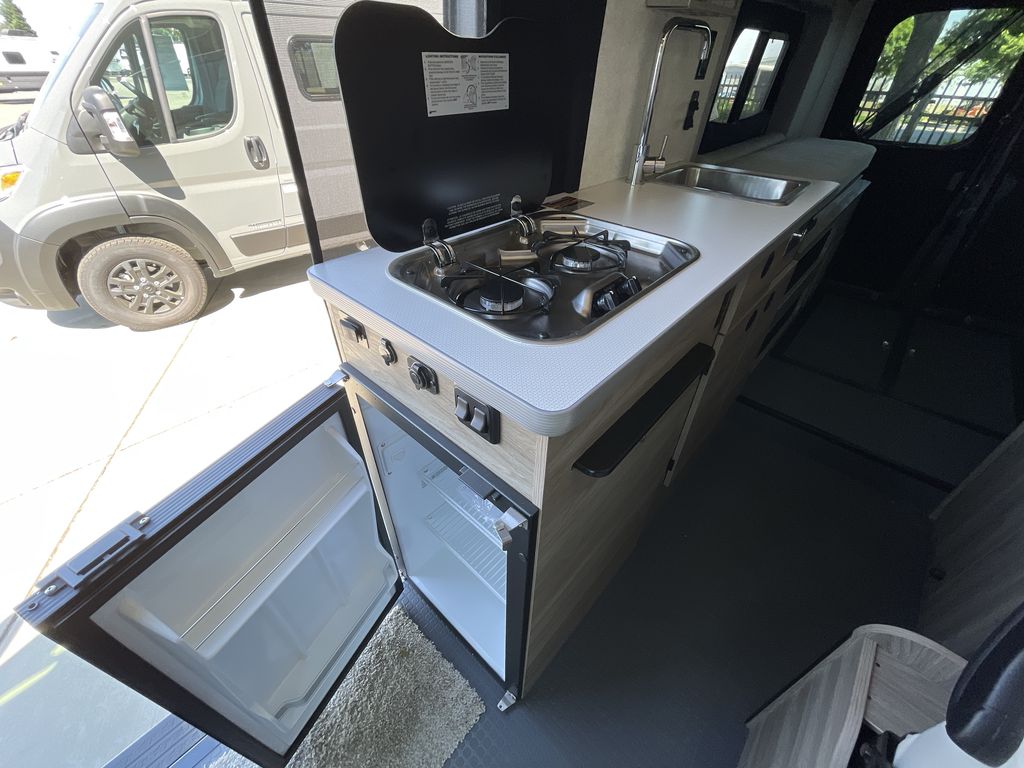  dry landscape.|RV rental camper van interior. Features light gray quilted passenger seats with seatbelts and a small dining table.|RV rental van kitchen: two-burner stove — photo 8