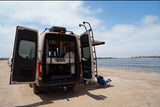 This RV rental is parked on a sandy beach with its rear doors open, revealing a bunk bed interior, ready for ocean adventures.