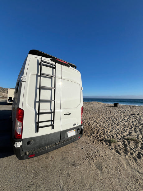 White RV rental van with a ladder, backed up to a sandy California beach. Clear blue ocean and sky complete the scenic view.