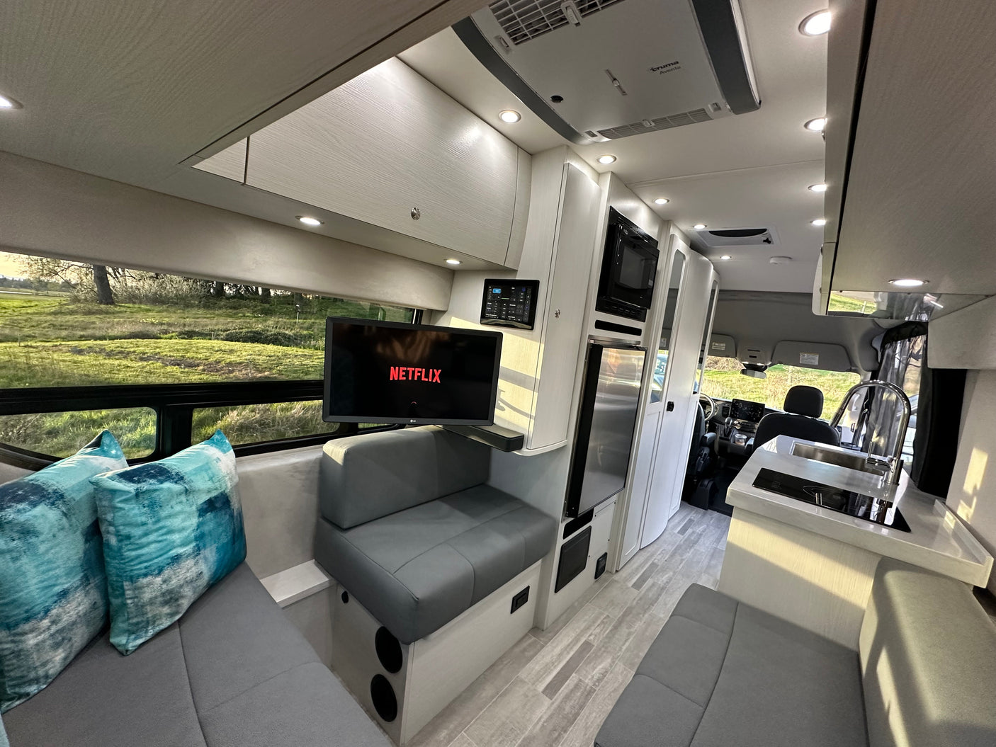 Step inside this luxurious RV rental, featuring a cozy living area with TV, modern kitchen, and scenic outdoor views.