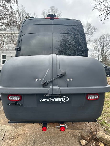 Rear of an RV rental van with a dark "Let's Go Aero" hitch-mounted storage box, showing integrated LED taillights.
