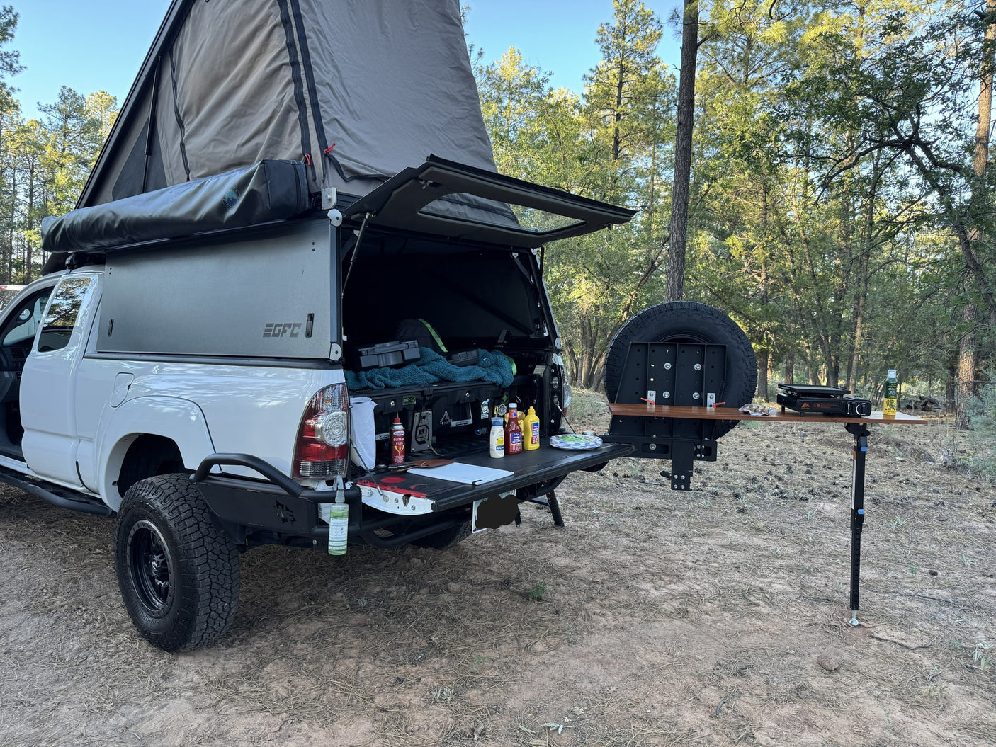 Overland-ready RV rental, white truck with pop-top camper & outdoor kitchen setup at a wooded campsite.