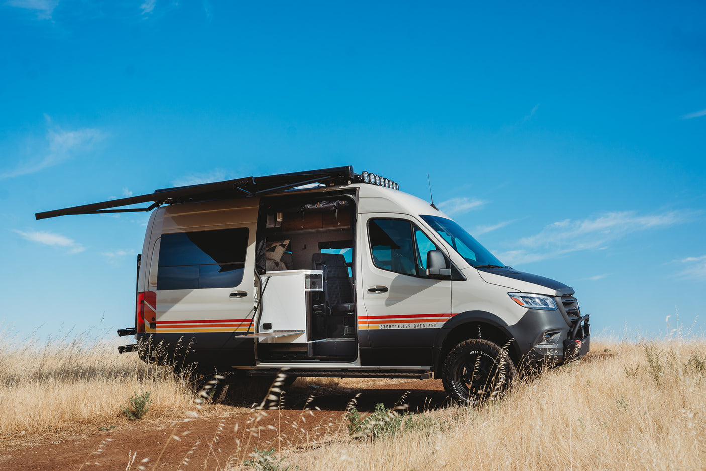 Experience van life! This Storyteller Overland camper van, ideal for an RV rental, is parked off-road with its side door open and awning out.