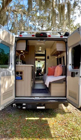 Back doors open on a cozy RV rental. Modern camper van interior features a kitchen, bed area, and path to the front cabin.