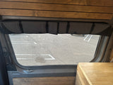 Inside an RV rental: window with a gray fabric insulating shade, wood panels, and a view of a parking lot.