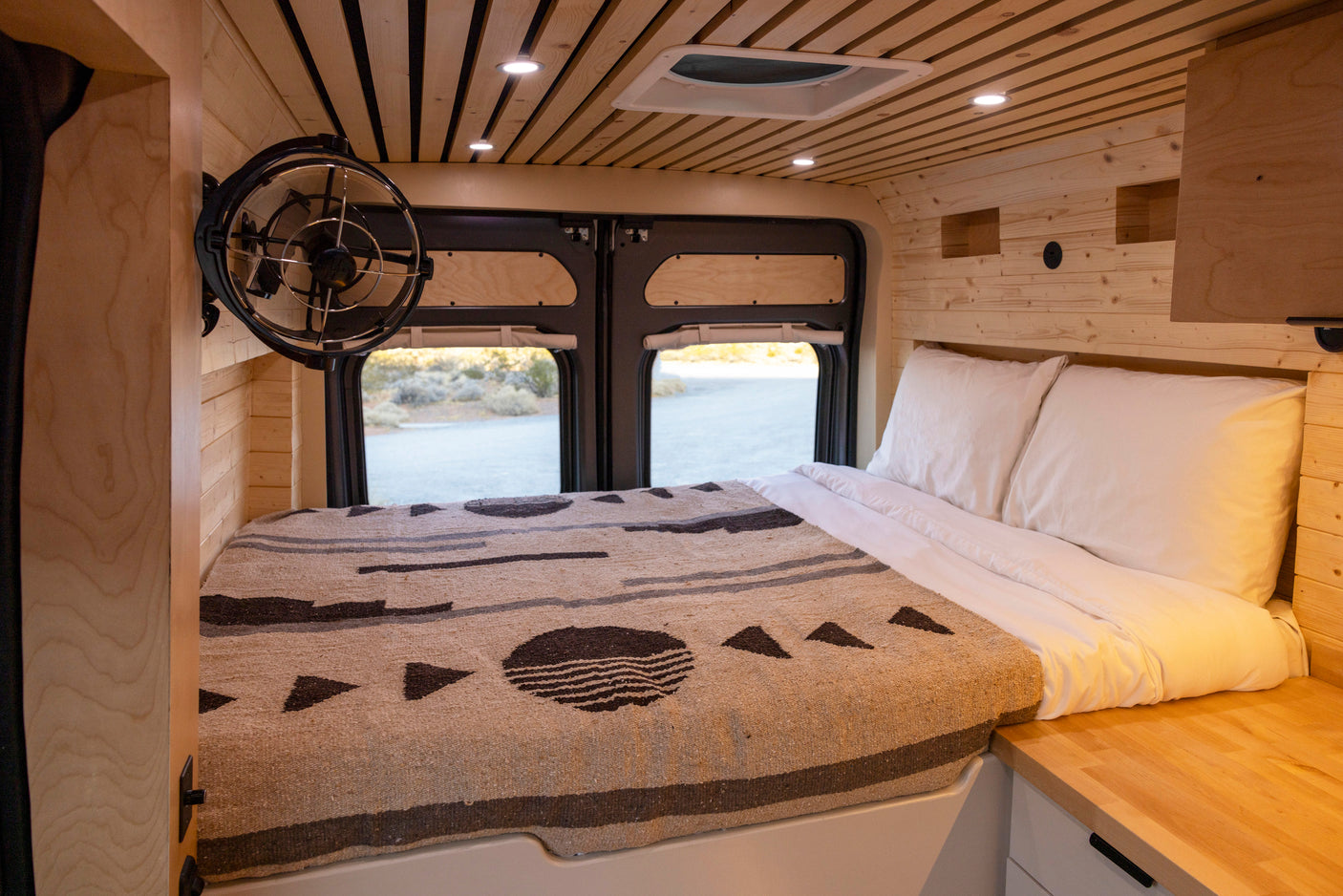 RV rental's inviting wood interior: bed, fan, and ceiling lights, with a view through rear windows.