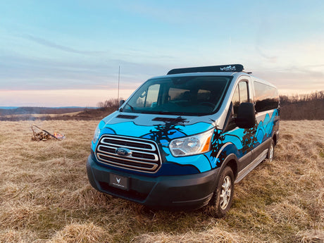 VanGo RV rental van, a customized Ford Transit with blue mountain art, parked in a scenic field with a campfire at dusk.