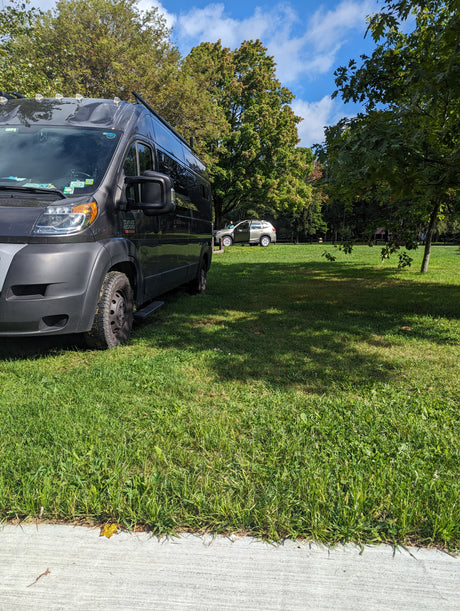 Experience nature with an RV rental! A dark gray camper van is parked on lush green grass next to a concrete path in a sunny park.