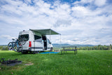 White RV rental camper van with bikes & awning, ready for adventure! Picnic table & fire pit in a scenic, grassy mountain field.