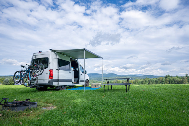 White RV rental camper van with bikes & awning, ready for adventure! Picnic table & fire pit in a scenic, grassy mountain field.