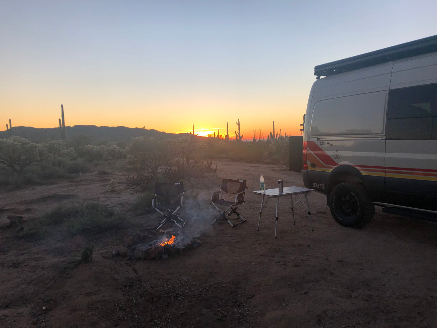 An RV rental parked in the desert at sunset, with a crackling campfire, chairs, and table ready for camping.