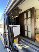 Inside a modern RV rental: open door view of a compact kitchen, fold-down table, stylish dark wood cabinets, and floor rug.