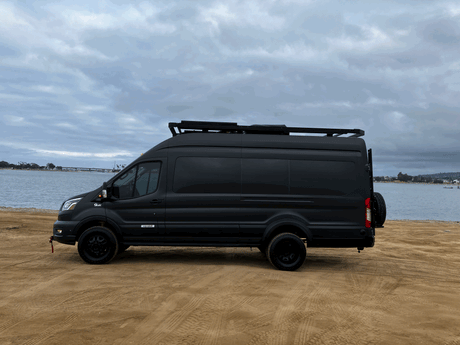 Black Vandoit camper van parked on sandy beach by water. Your next RV rental adventure awaits!