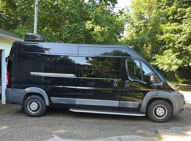 Side view of a black camper van with a roof AC unit, parked on asphalt. Consider this modern RV rental for travel.