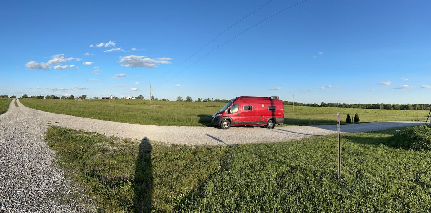 A red RV rental is parked on a winding gravel road in a vast green field under a clear blue sky, inviting exploration and road trips.