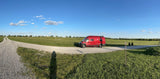 A red RV rental is parked on a winding gravel road in a vast green field under a clear blue sky, inviting exploration and road trips.