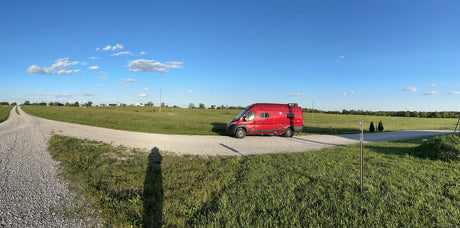 A red RV rental is parked on a winding gravel road in a vast green field under a clear blue sky, inviting exploration and road trips.