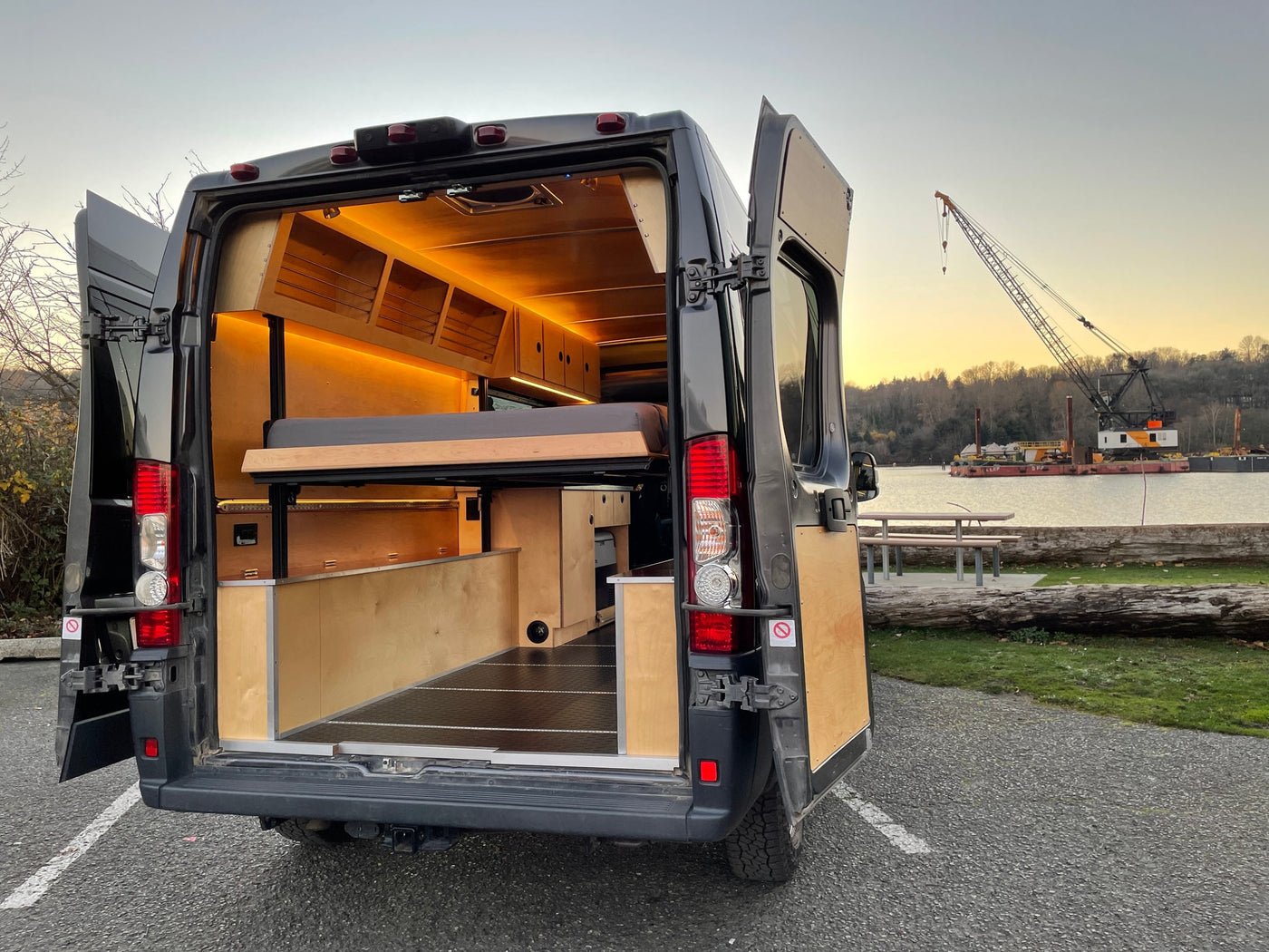 Explore this modern RV rental's rear, open doors showcasing a warm, lit wood interior with a raised bed & storage. Parked by water. — photo 4
