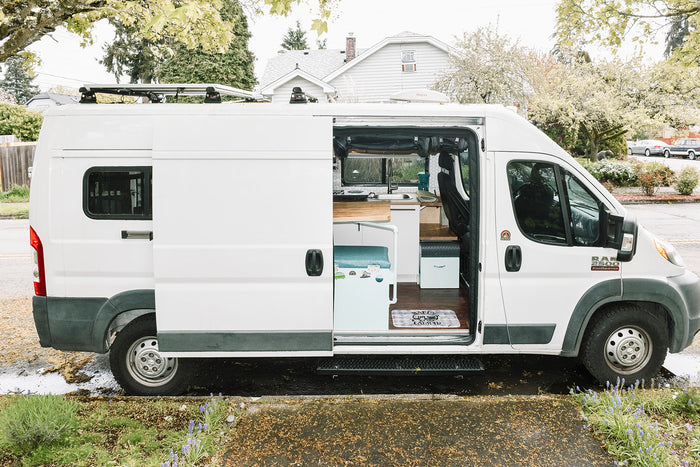 This white Ram ProMaster converted camper van, ideal for rv rental, has its side door open, revealing a compact kitchen and living area inside.