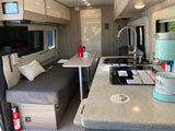 RV rental interior: compact kitchen with sink & stovetop, a coffee station, TV, and bench seating.