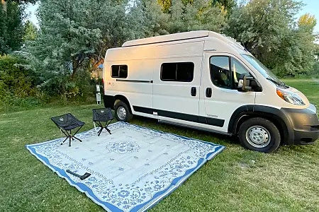RV rental ready for adventure: white camper van parked with outdoor rug & chairs on grass.|White Ram ProMaster camper van with pop-top roof for RV rental