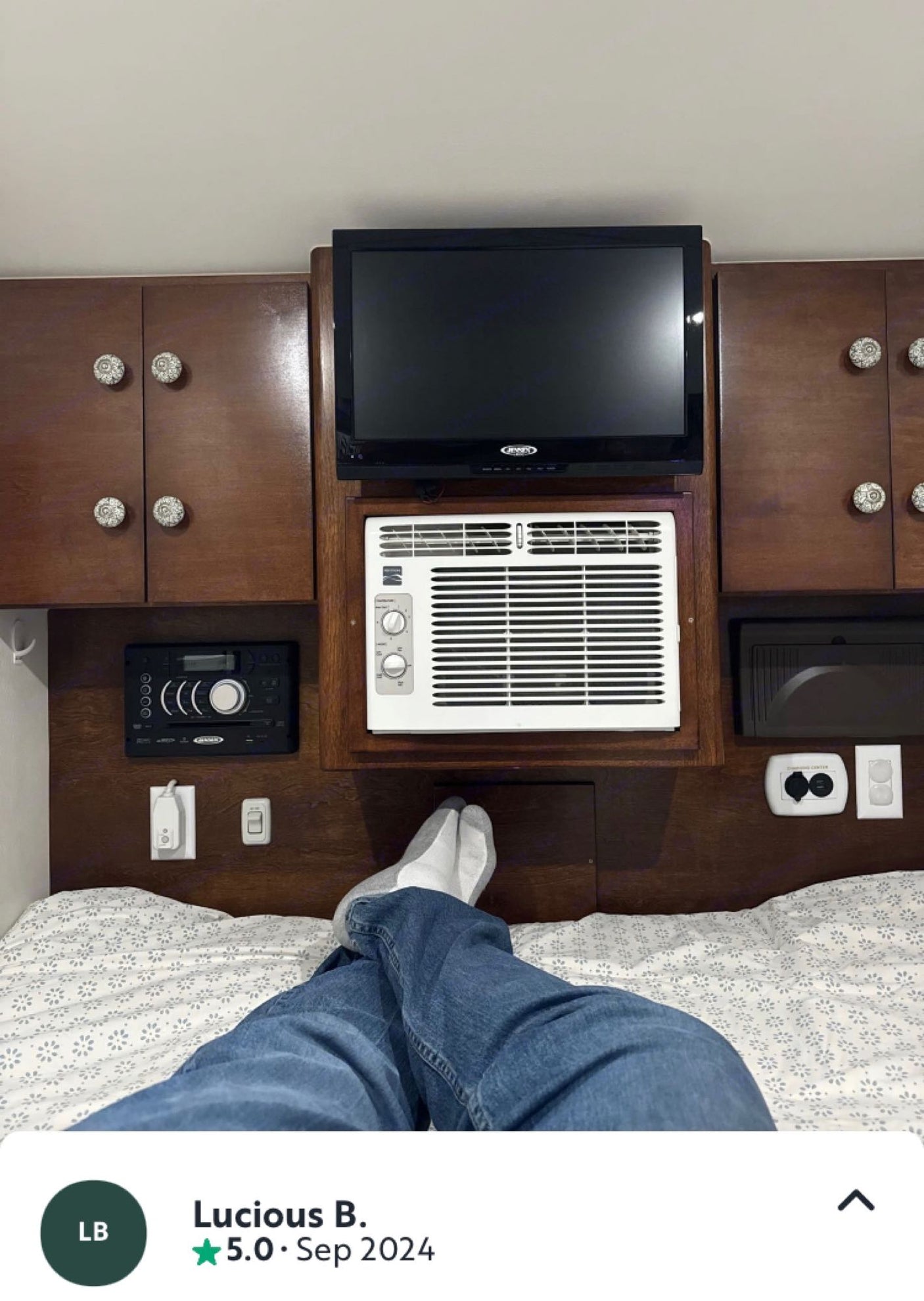 Relaxing in an rv rental: Person's legs on bed, TV, AC, stereo, & charging station built into wooden panel above.