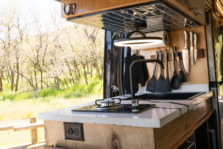 Modern RV rental kitchen setup with a stovetop, sink, hanging utensils, and a scenic outdoor view.