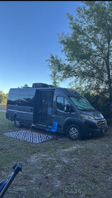Our grey RV rental parked in a grassy spot. The campervan's side door is open, with a rug and blue chair set up for outdoor relaxation.