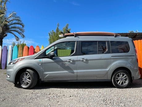 Compact grey van with surfboard, perfect for a beach rv rental trip. Parked by colorful surfboards under a blue sky.