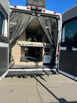 Inside an RV rental camper van: rear view shows a bed, storage, and utility panel, all through a protective mesh screen.
