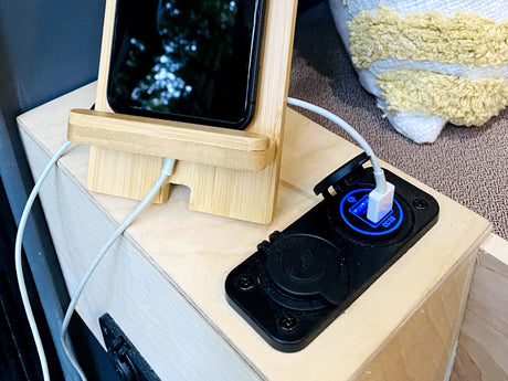 An RV rental interior: a smartphone on a bamboo stand charges via a blue-lit 12V USB port on a light wood surface.