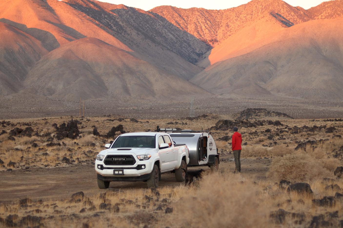 White Toyota truck towing an RV rental teardrop trailer in a vast desert with sunlit mountains. A person and dog are outside.