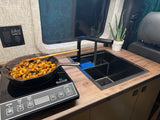 RV rental kitchen scene: Pasta cooking on an induction cooktop. Features a modern sink, counter, and power outlets.